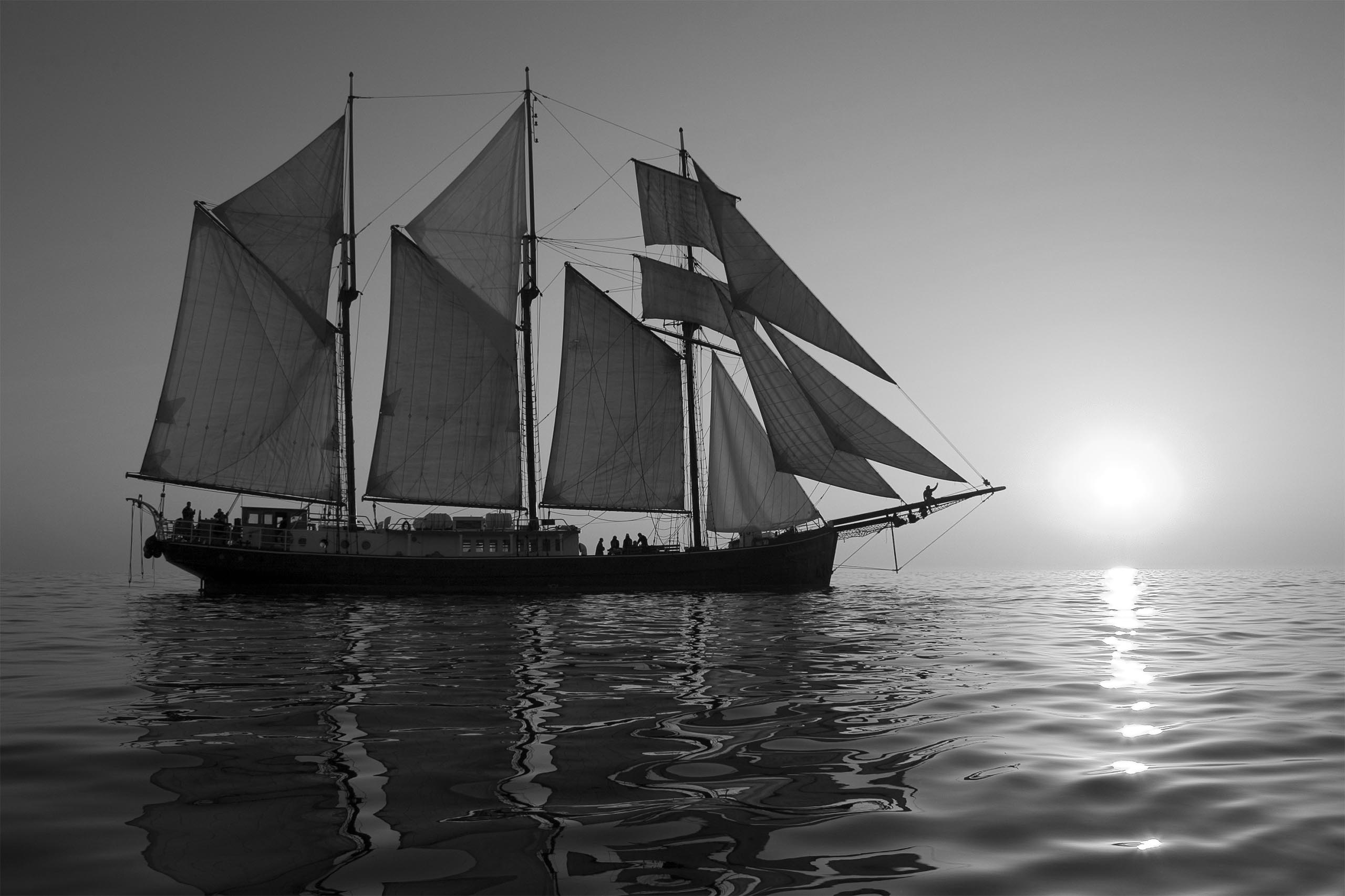 Black and white photo of three-masted schooner with sun setting on the horizon