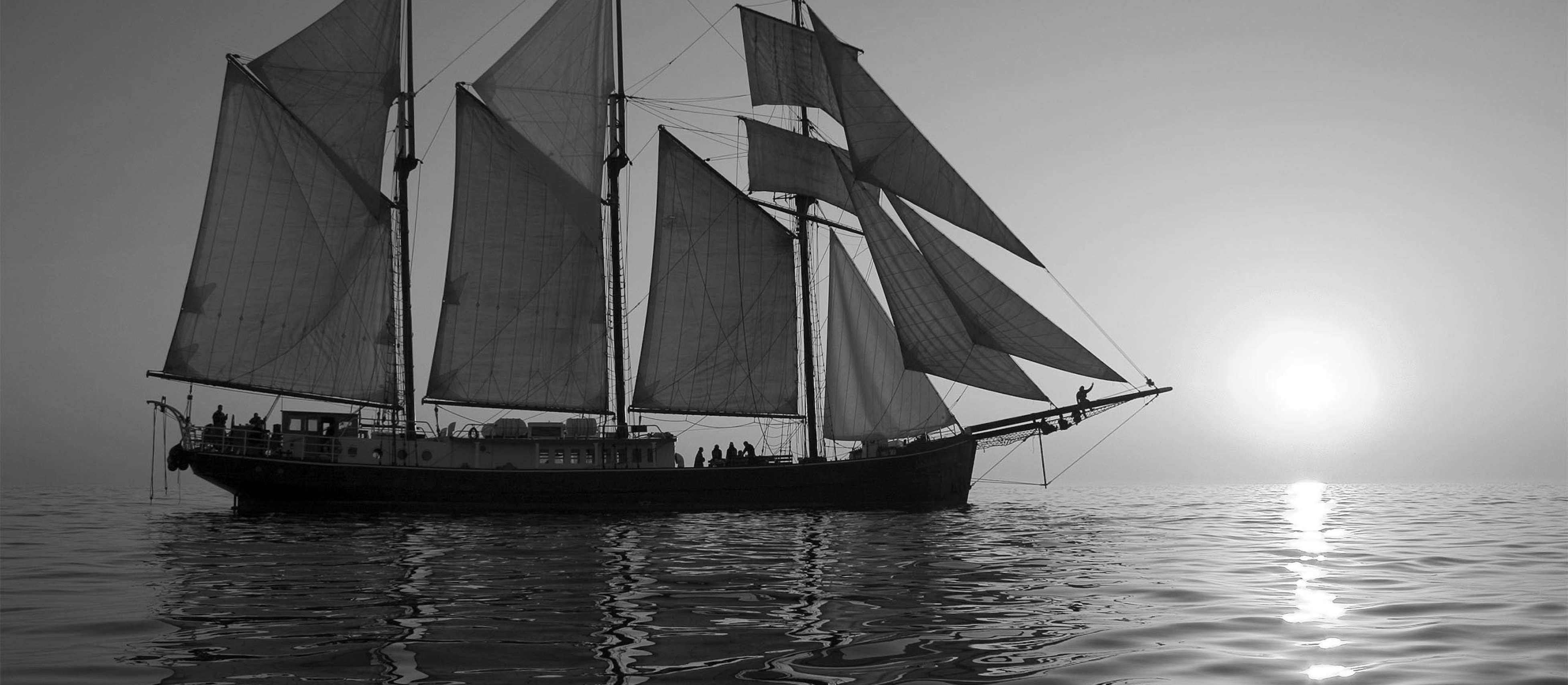Black and white photo of three-masted schooner with sun setting on the horizon